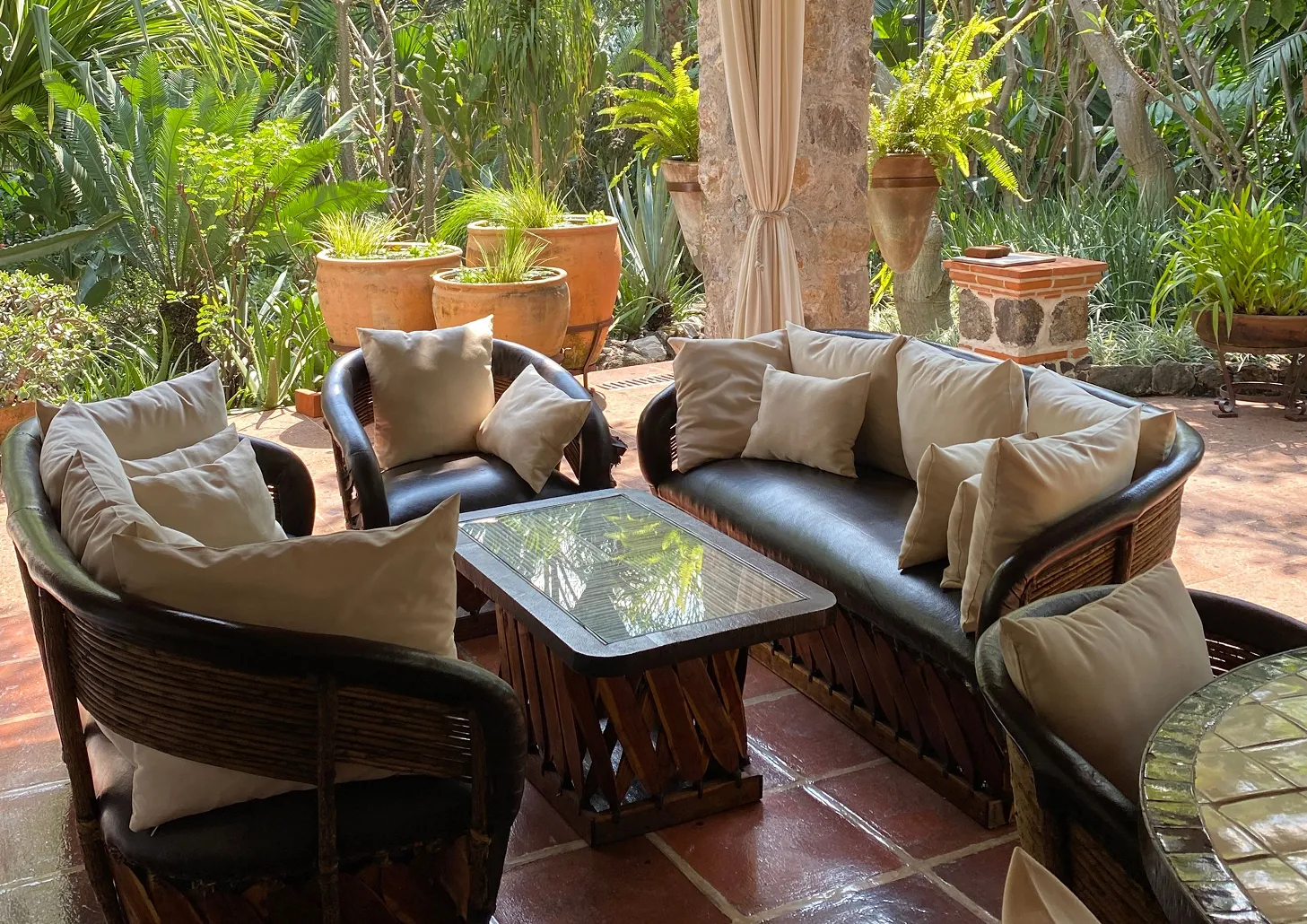 Outdoor seating area with dark wicker chairs and sofa featuring beige cushions around a glass-top wooden coffee table, surrounded by green plants and terracotta pots.