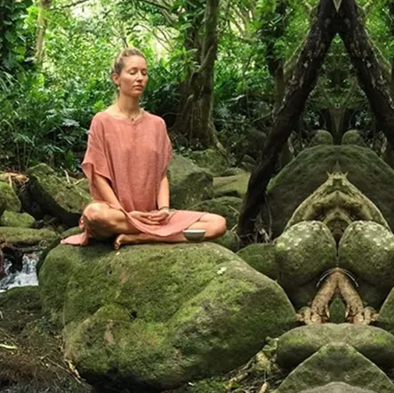 Woman in a pink dress meditating cross-legged on a moss-covered rock in a lush green forest.