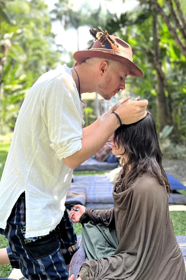 Man in white shirt and hat with feathers performing a ritual or healing on a seated woman outdoors.