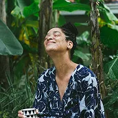 Smiling woman with braided hair in a leafy outdoor setting holding a beverage cup.