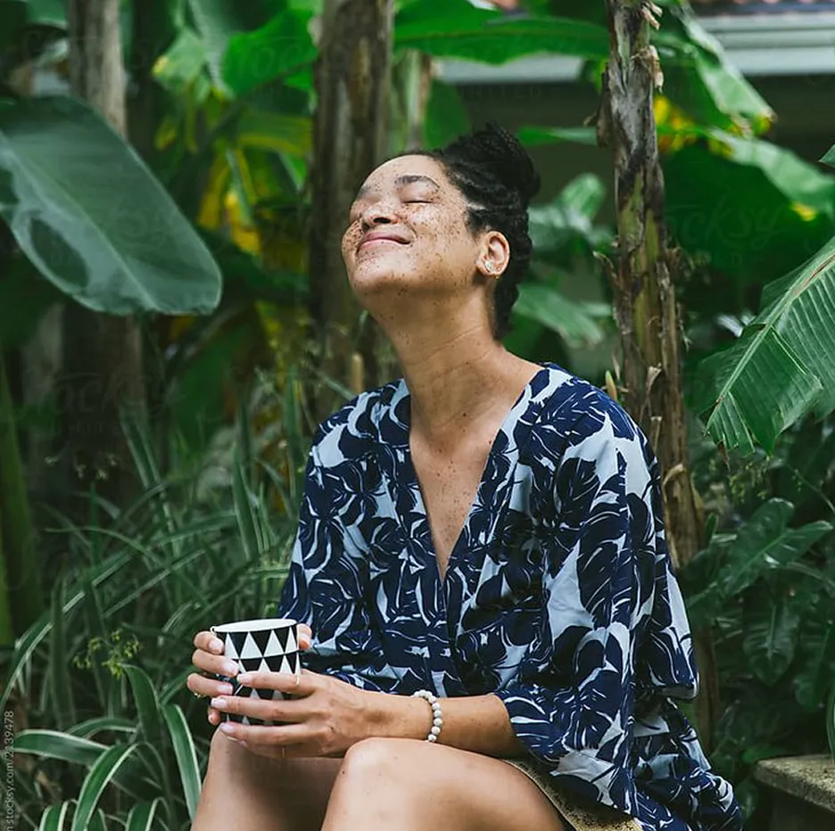 Woman with freckles sitting outdoors among green plants, smiling with eyes closed while holding a black and white patterned cup.