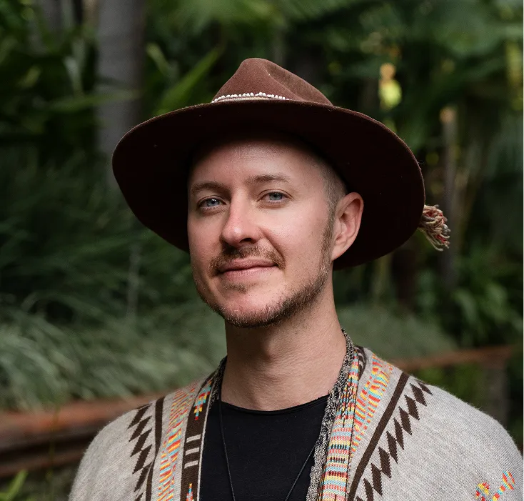 Smiling man wearing a brown hat and colorful patterned sweater with green foliage in the background.