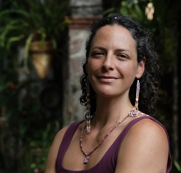 Smiling woman with curly dark hair wearing long beaded earrings and a purple sleeveless top, standing outdoors with greenery in the background.