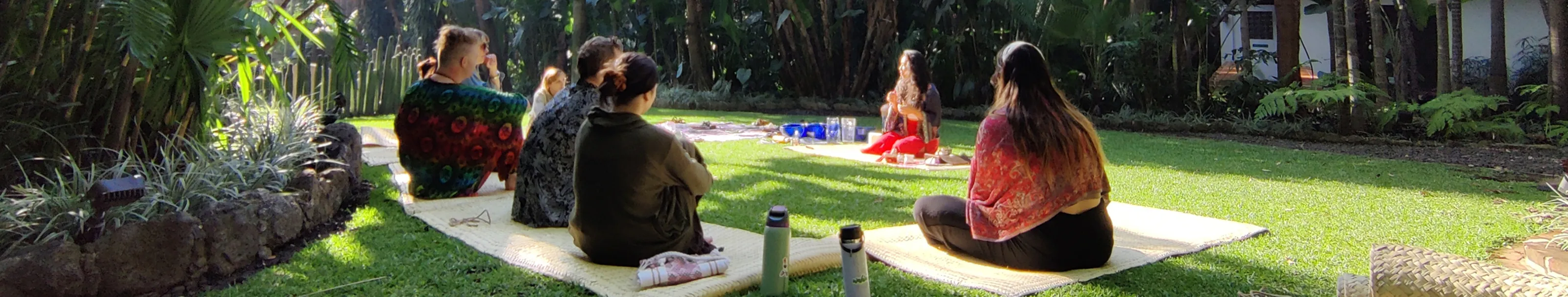 Group of people sitting on mats on grass in a garden, facing a person speaking during a sunny outdoor gathering.