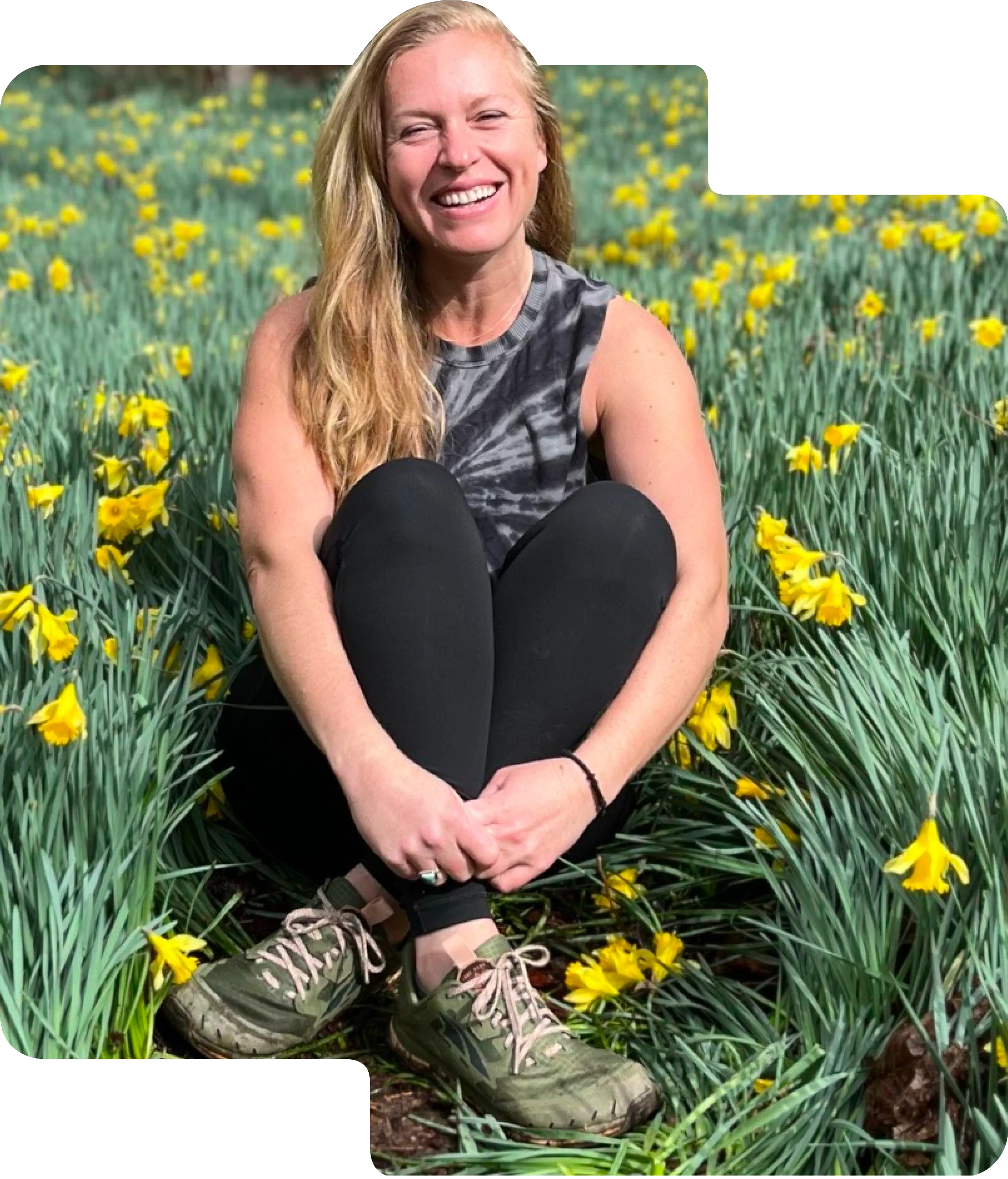 Smiling woman with long blonde hair, wearing a sleeveless tie-dye shirt and black leggings, sitting cross-legged in a field of yellow daffodils.