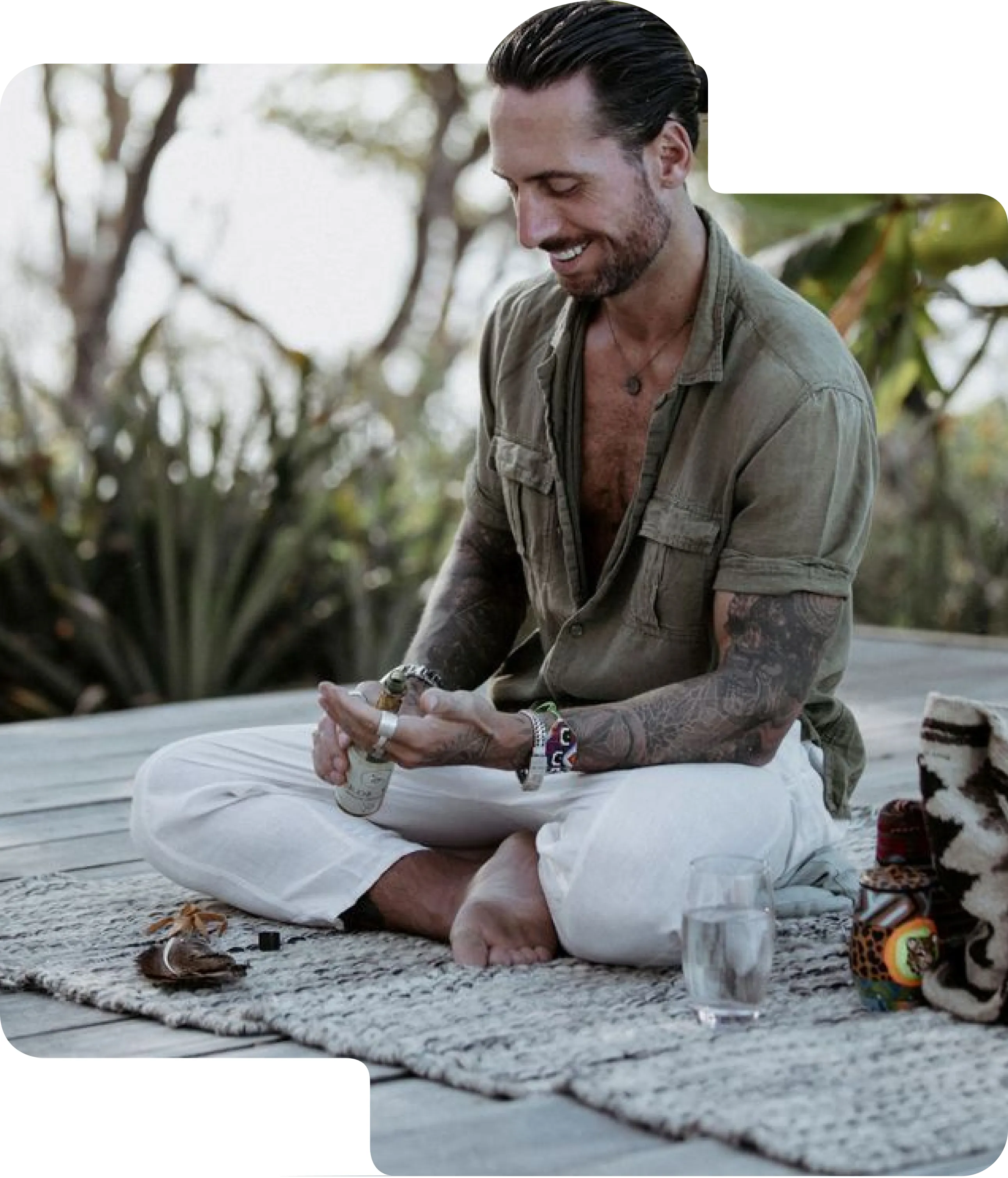 Man with tattoos sitting cross-legged on a rug outdoors, smiling while applying spray to his hands.