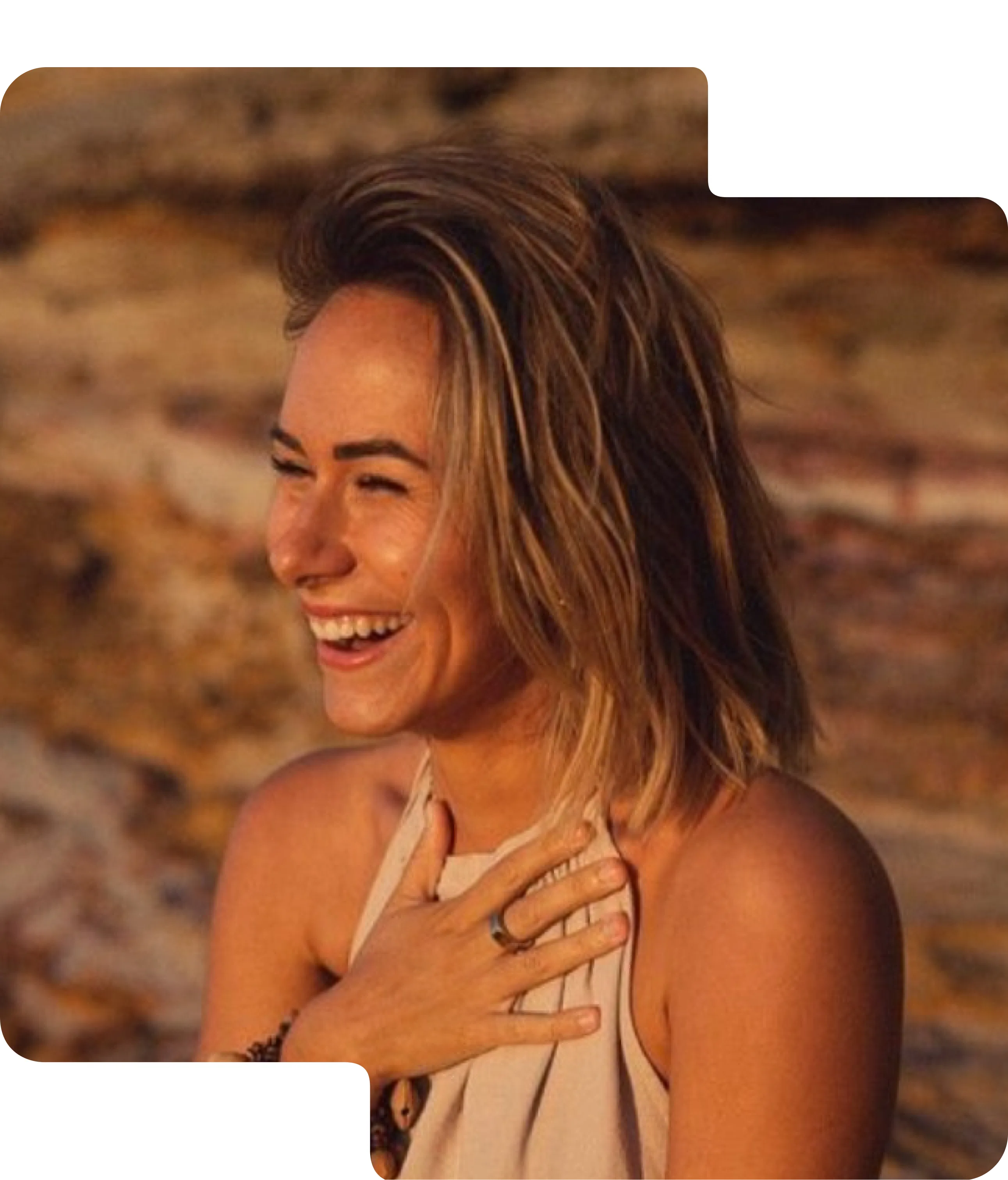 Smiling young woman with shoulder-length blonde hair, wearing a sleeveless top, holding her hand to her chest outdoors.