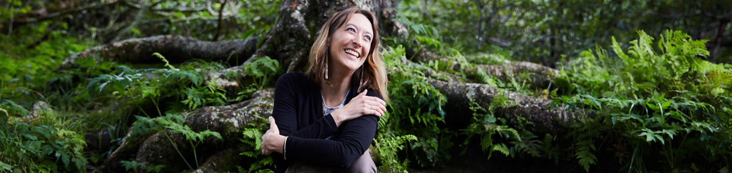 Smiling woman sitting in front of large tree roots surrounded by green foliage in a forest.