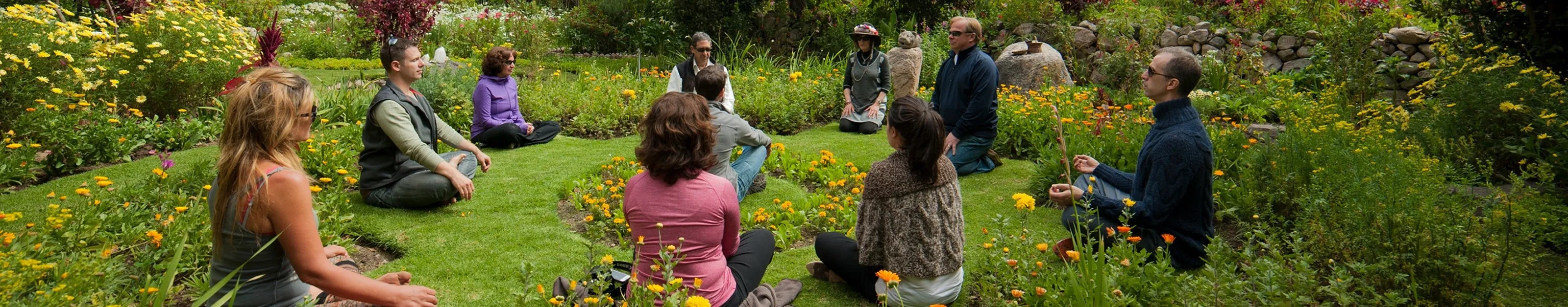 Group of people meditating in a circle on grass surrounded by a flower garden with yellow and orange blooms.