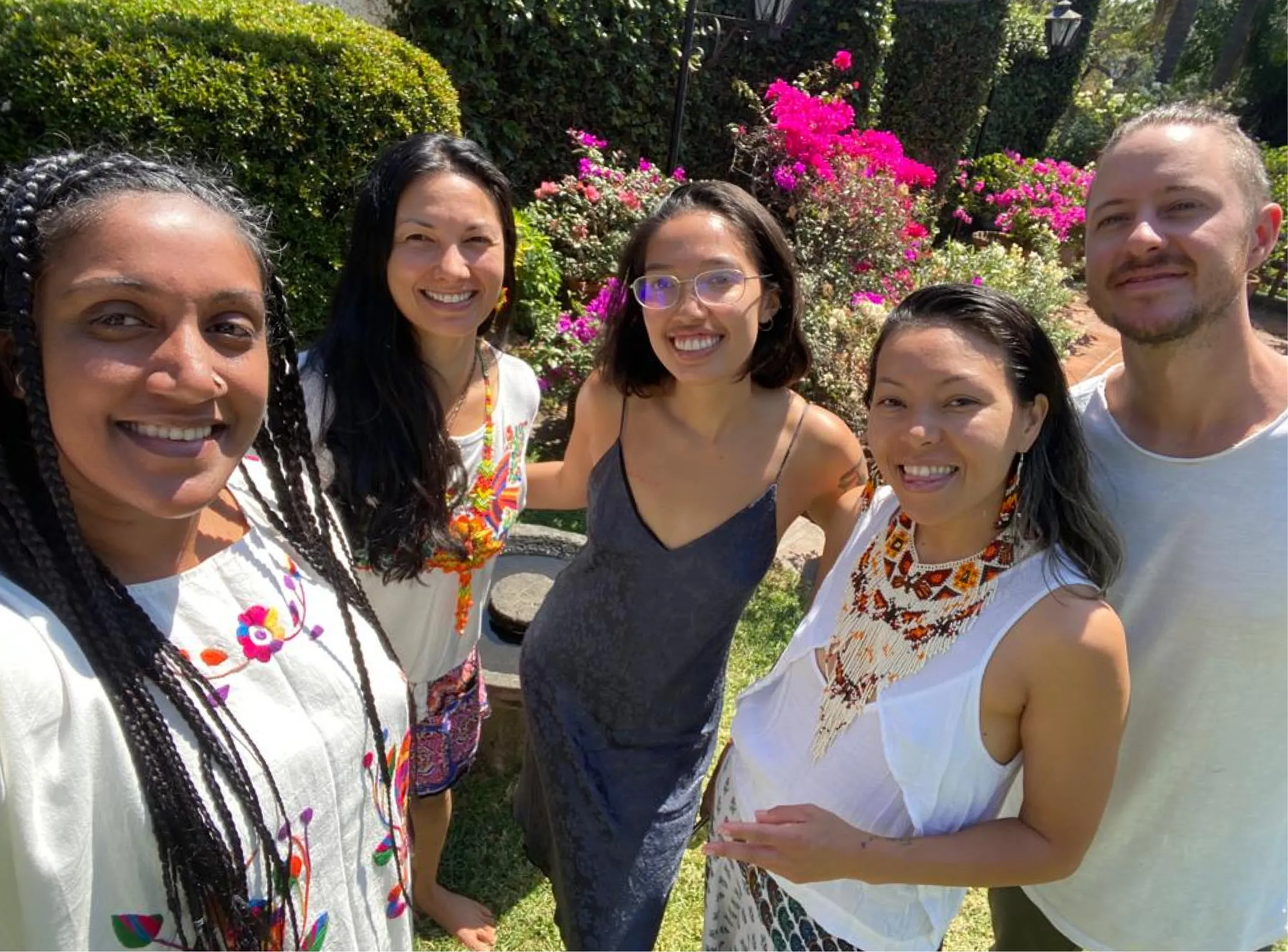 Five smiling people posing outdoors in a garden with colorful flowers and greenery in the background.
