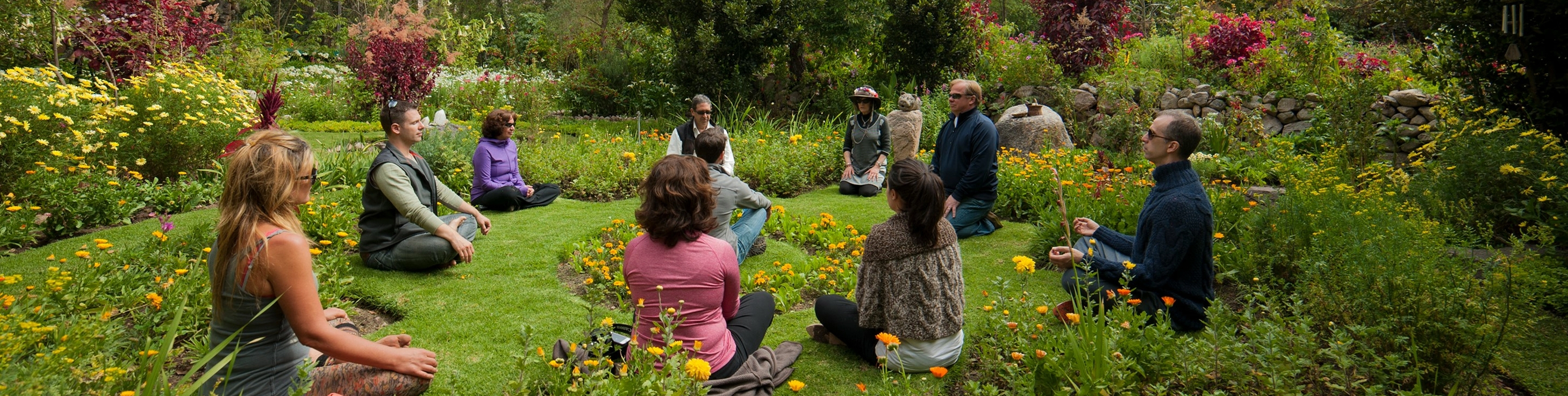 Group of people sitting cross-legged in a circle on grass surrounded by colorful flowers in a garden.