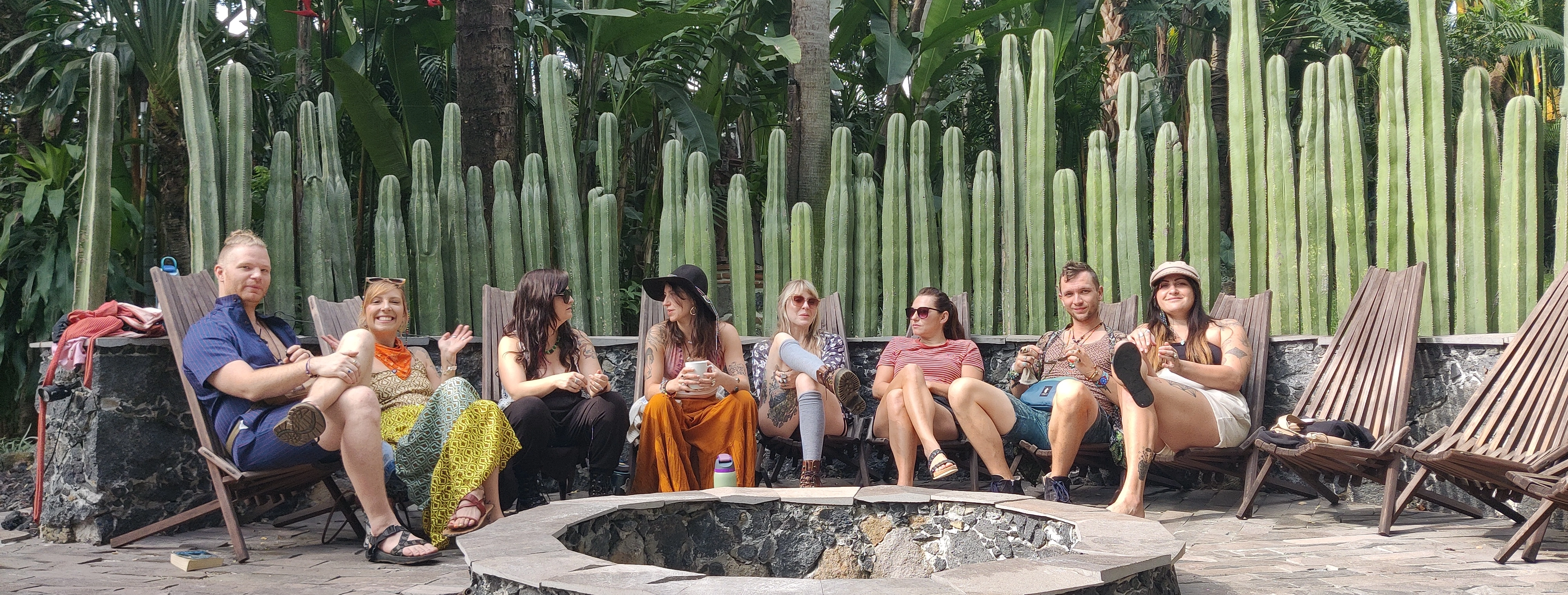 Group of eight people sitting on wooden chairs around a stone fire pit with tall cactus plants and tropical greenery in the background.