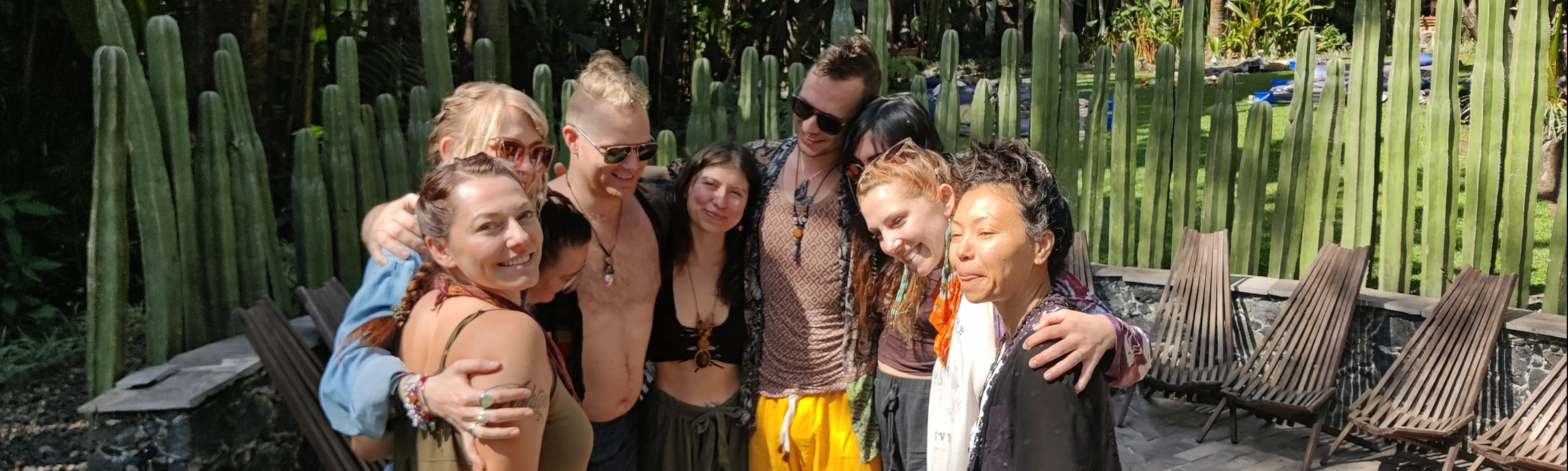 Group of eight smiling young adults with arms around each other standing outside near tall cacti and wooden chairs.