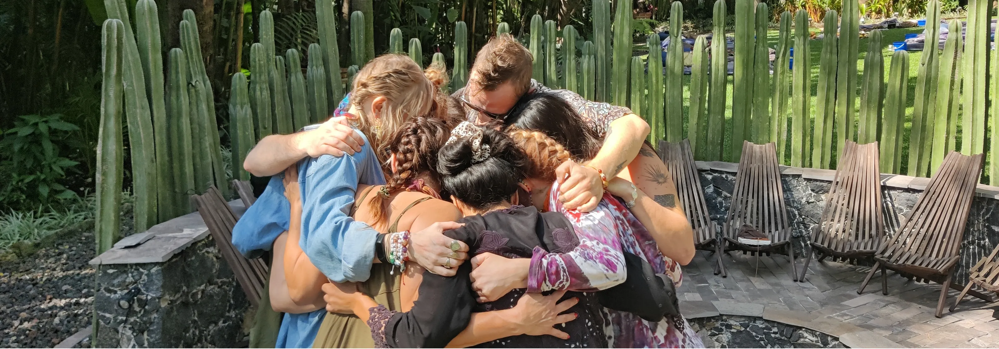Group of six people in a close group hug outdoors near tall cacti and wooden chairs.