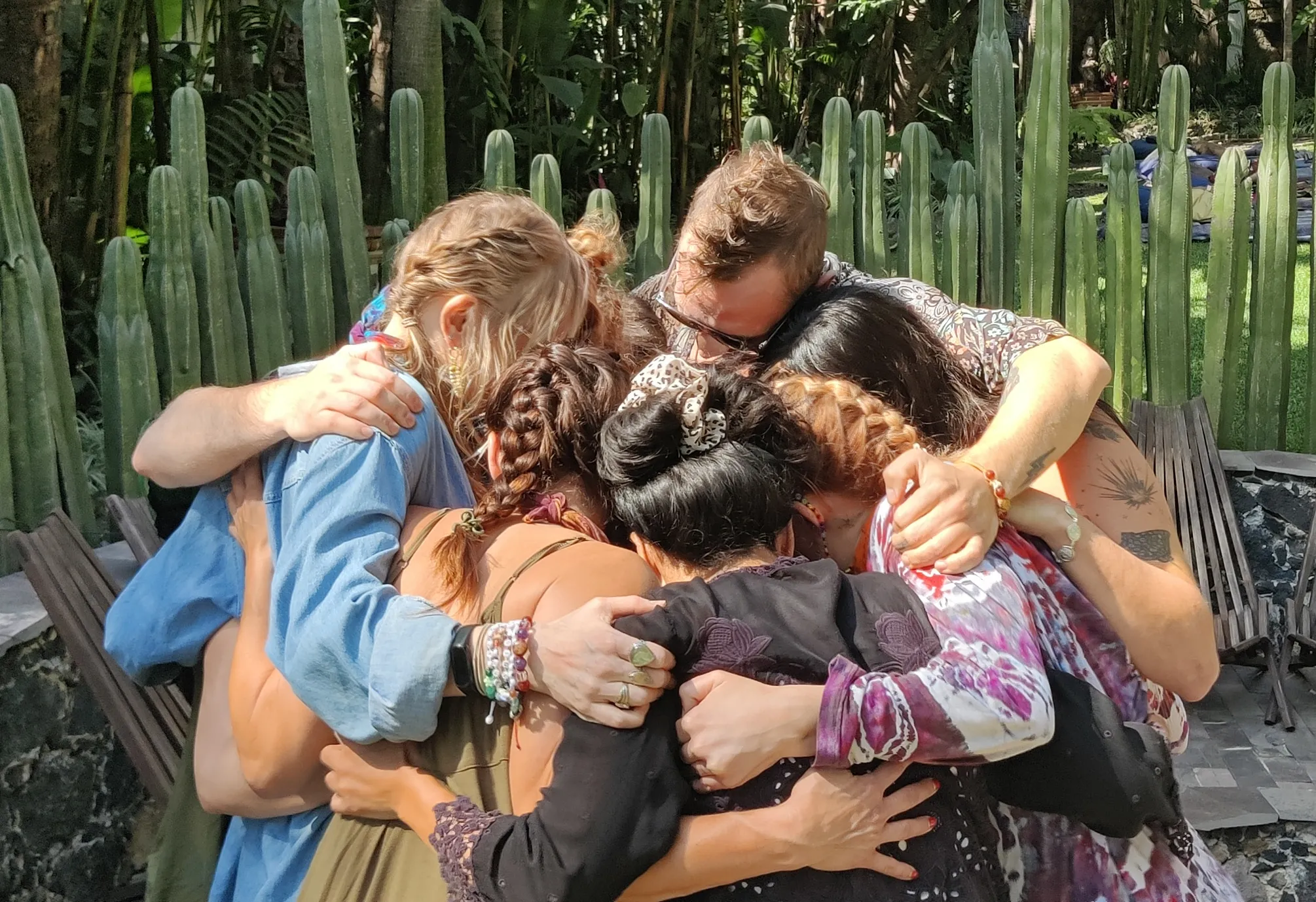 Group of six people tightly embracing in a circle outdoors near tall cacti.