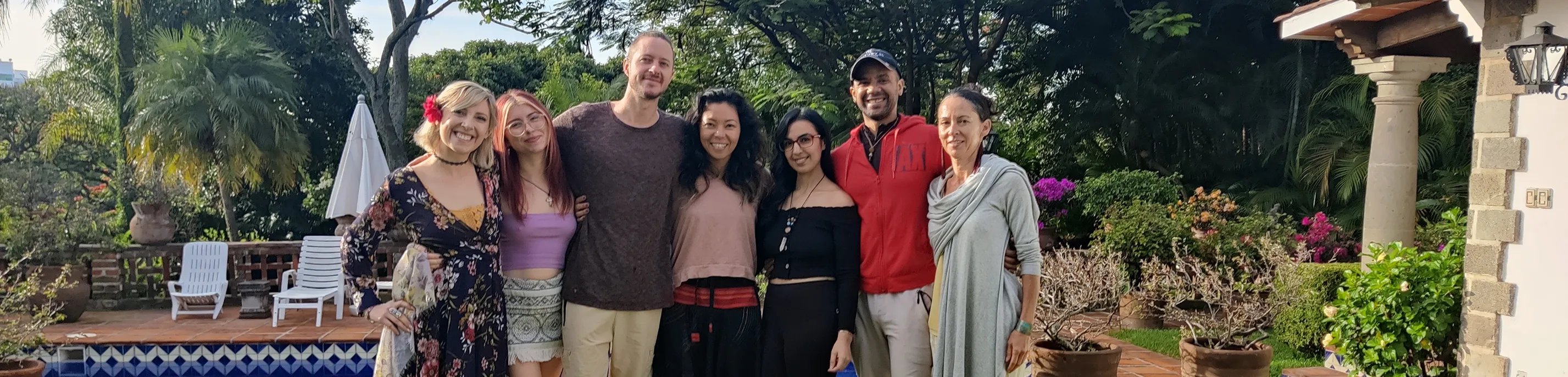 Seven people standing side by side smiling outdoors near a pool with garden and trees in the background.