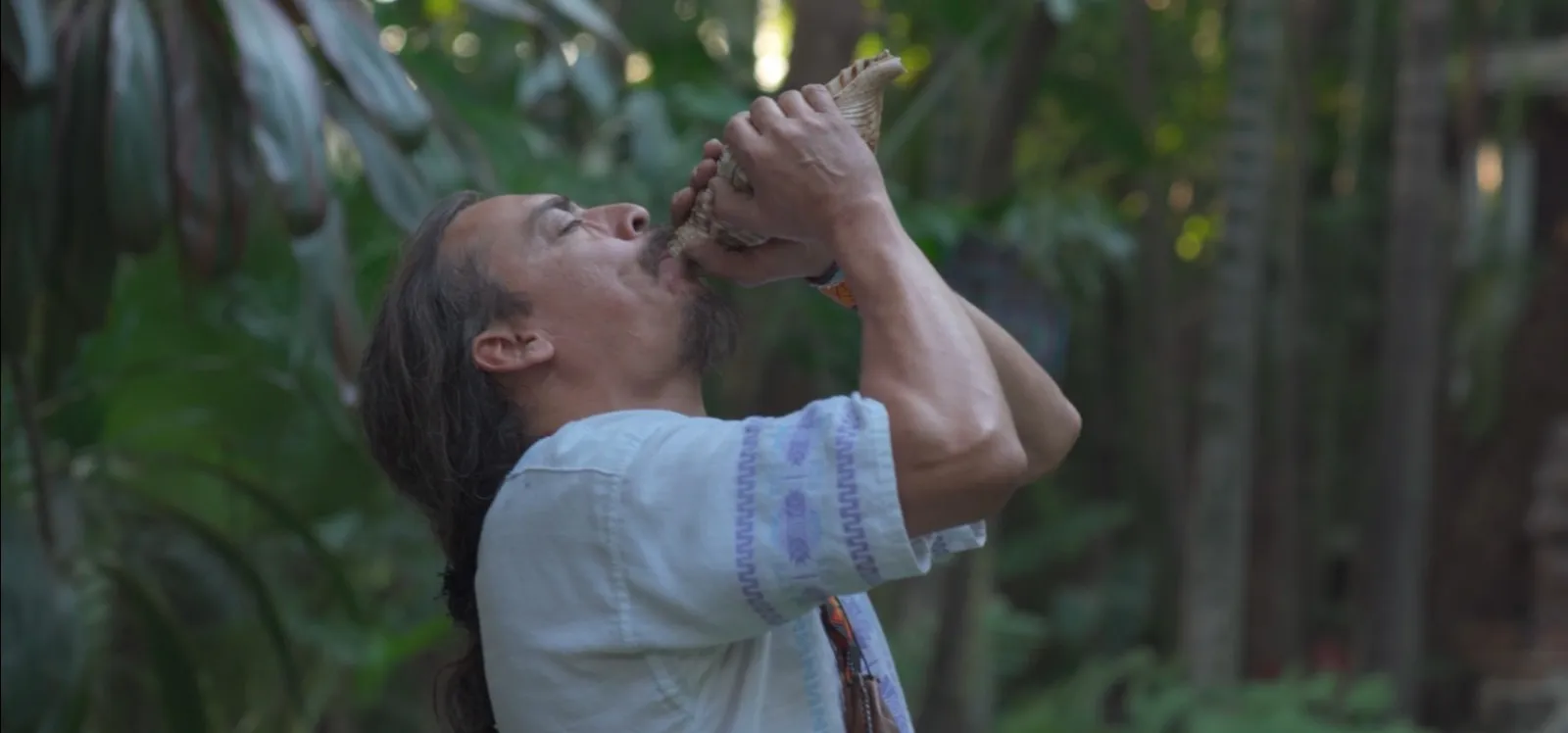 Man blowing into a conch shell in a lush outdoor setting with green foliage.