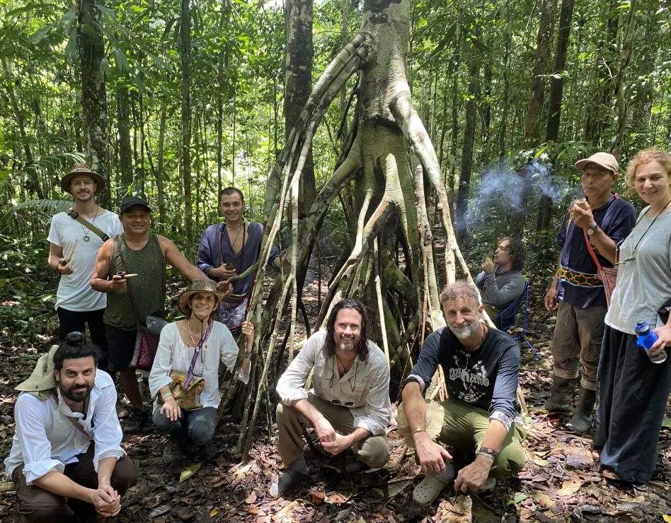 Group of people posing around a large tree with exposed roots in a dense forest, some seated and others standing, with one person smoking.