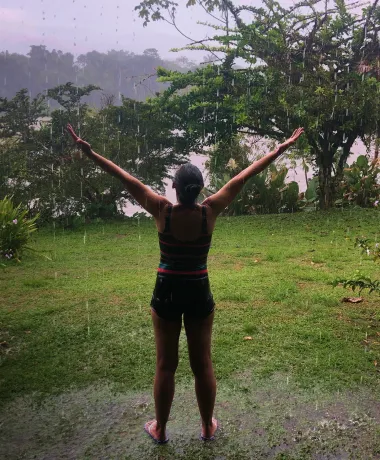 Woman in athletic wear standing barefoot on wet grass with arms raised, facing lush greenery in the rain.