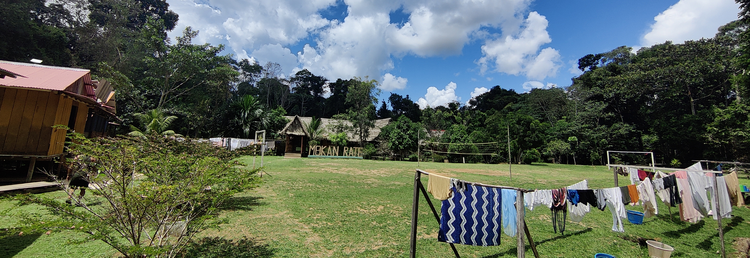 Clothes hanging on lines to dry in a grassy area with wooden buildings, volleyball net, and lush green trees under a partly cloudy sky.