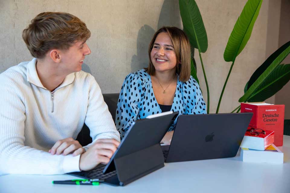 Zwei Mitarbeiter im Gespräch über steuerrechtliche Inhalte mit Laptops und Gesetzbüchern am Tisch