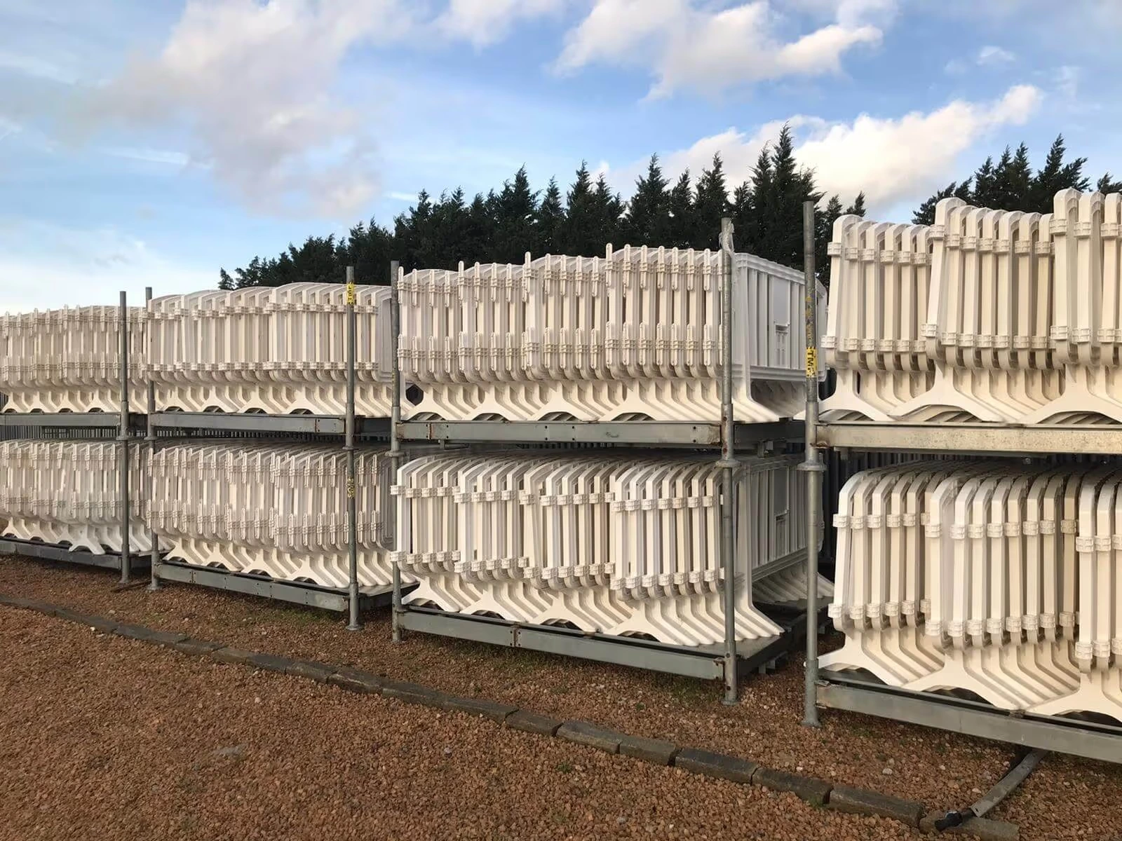 Racks de stockage contenant des barrières de police en plastique blanc empilées, installés en extérieur sur sol gravillonné.
