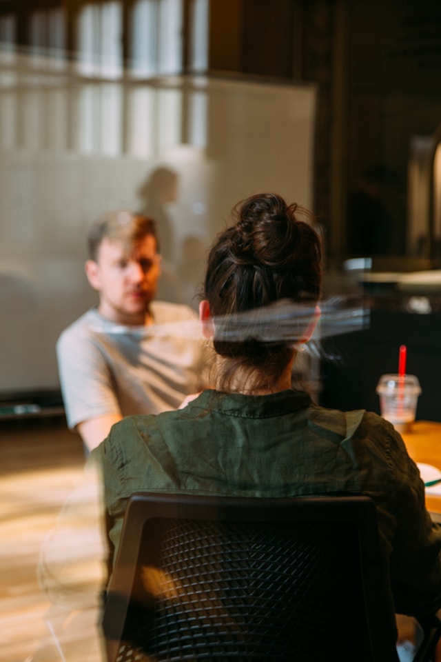 Two people having a discussion in an office setting, viewed through a glass partition with reflections.