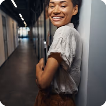 A person leaning against the wall of an office, holding a laptop.