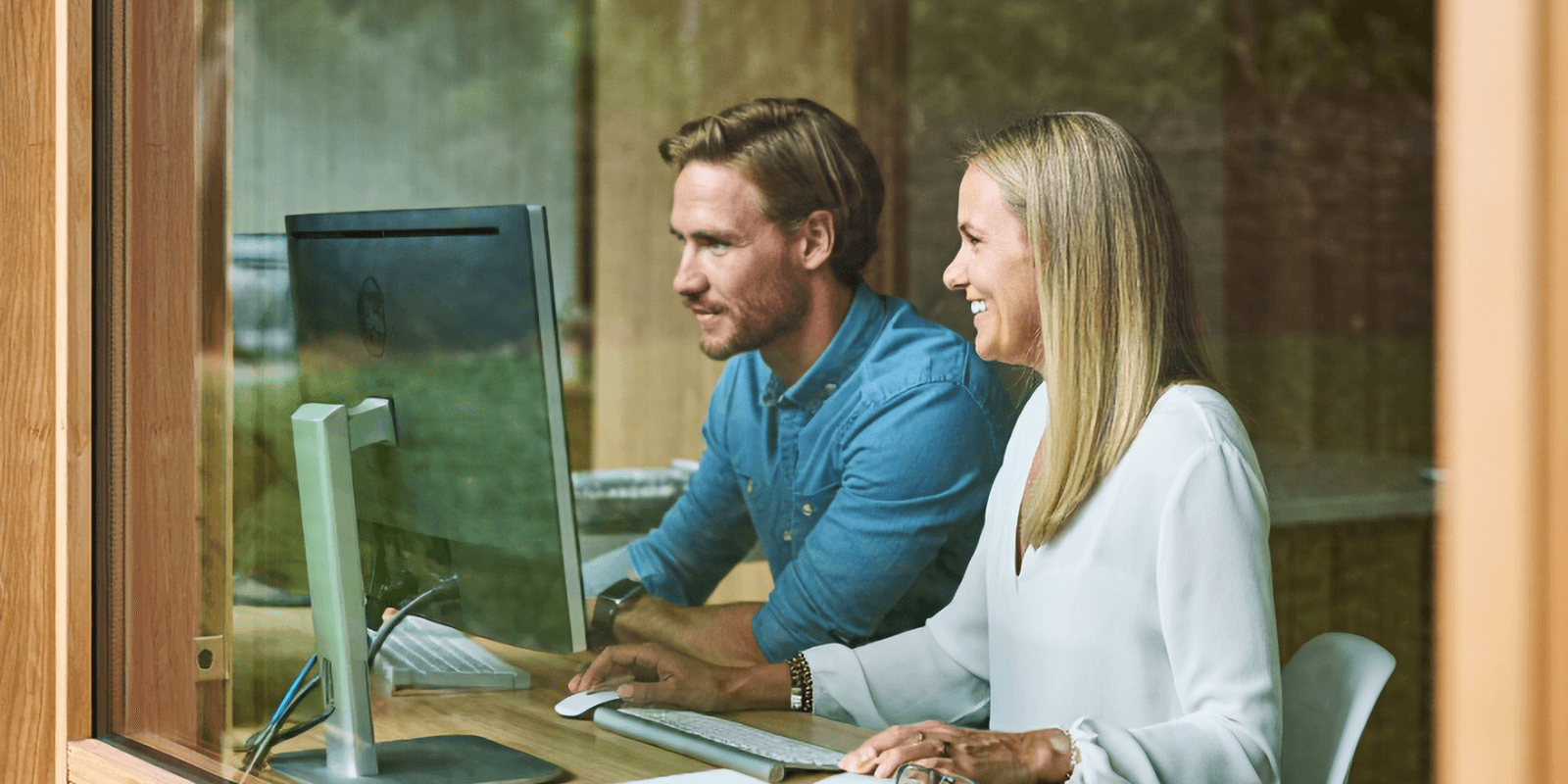 Blick durch ein Fenster auf einen Mann in einem blauen Hemd und eine Frau mit blonden Haaren in einem weißen Pullover. Die beiden sitzen an einem Schreibtisch und blicken gemeinsam lächelnd auf einen großen Computer-Bildschirm.