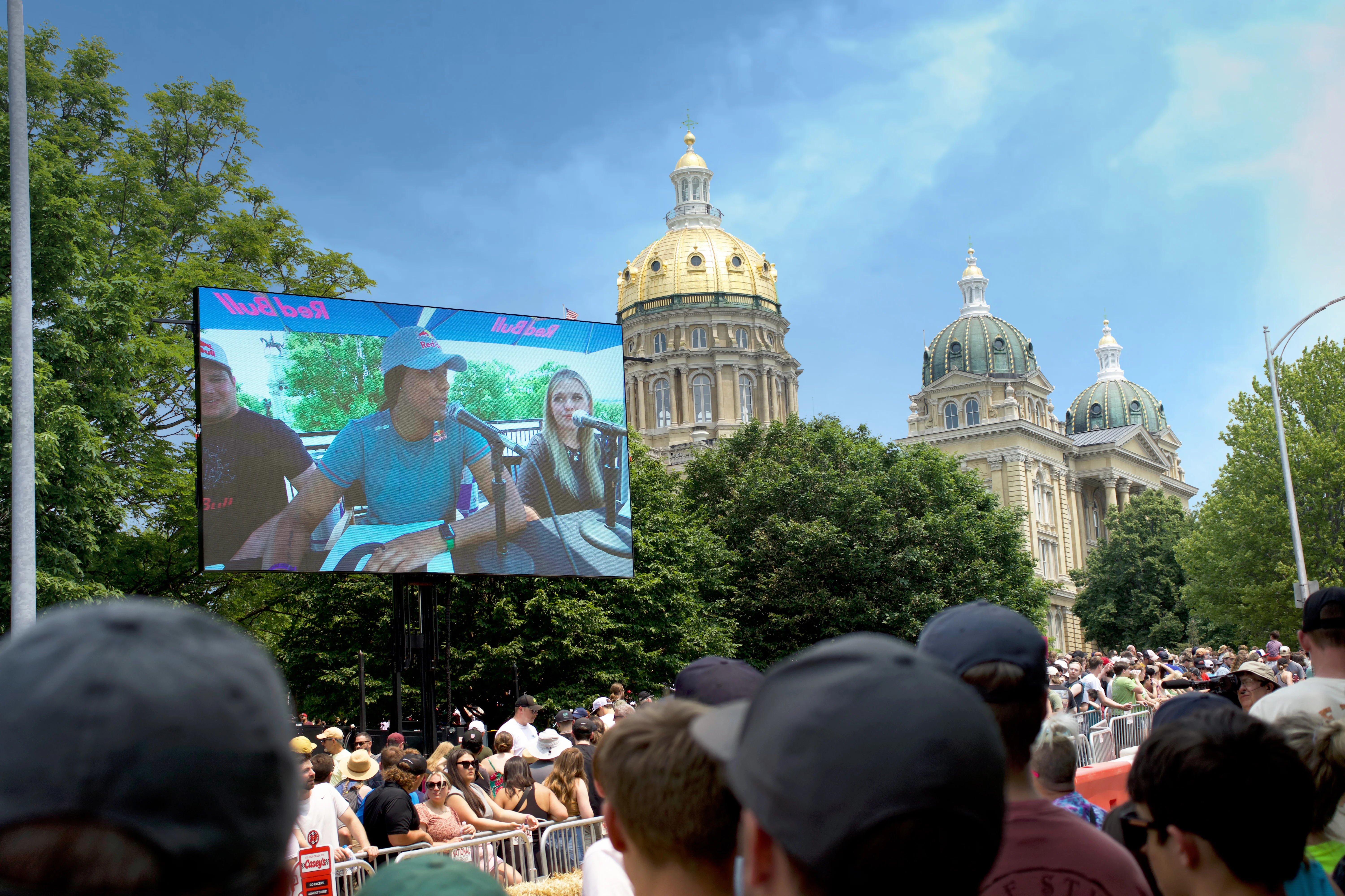 A large crowd watches a panel of judges on a mobile LED screen at the Red Bull Soapbox Race, with the Iowa State Capitol’s golden dome and green-roofed towers prominently visible behind the trees.