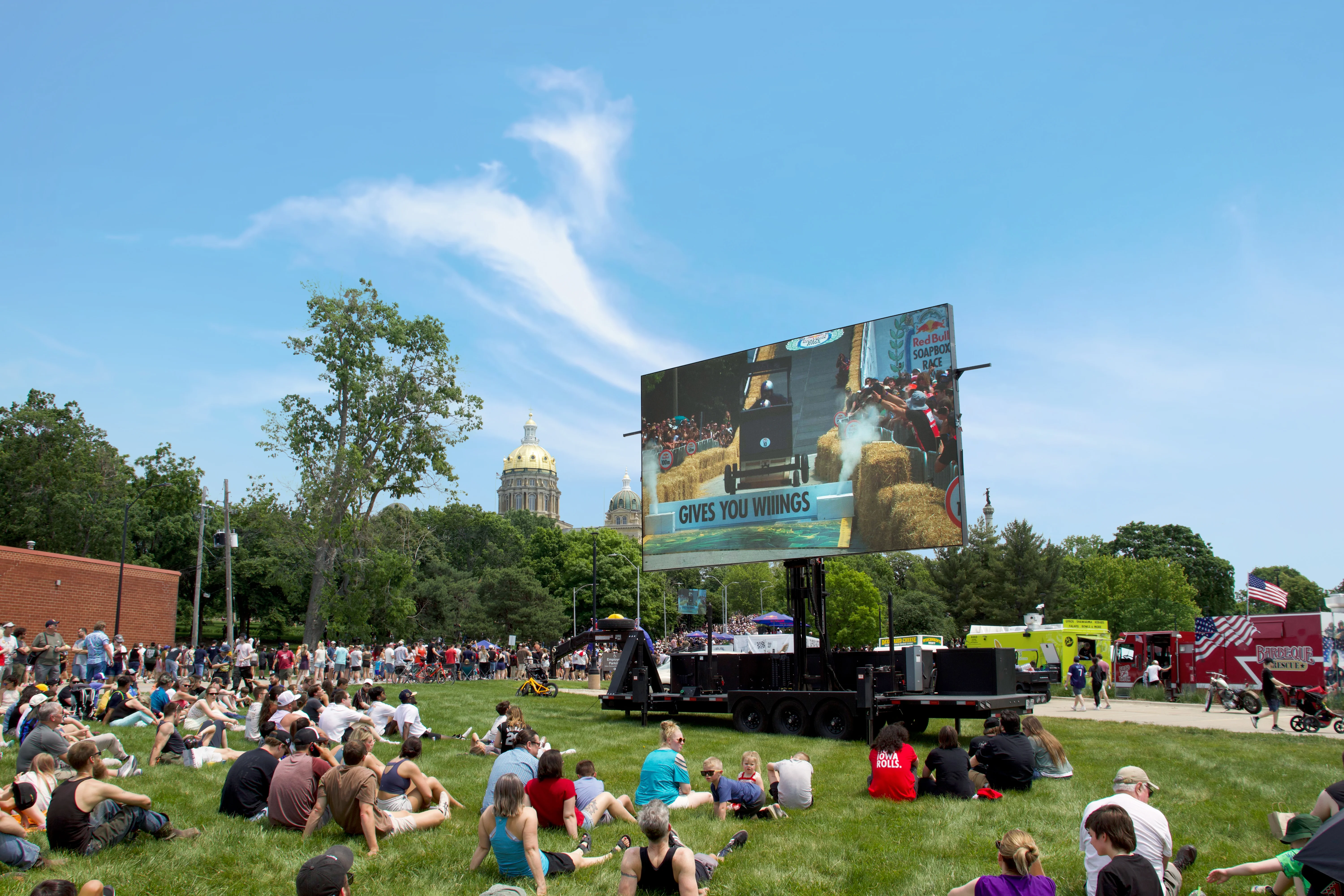  Spectators sit on a grassy lawn watching a large mobile LED screen displaying a soapbox car racing down a track. The golden dome of the Iowa State Capitol rises in the background.