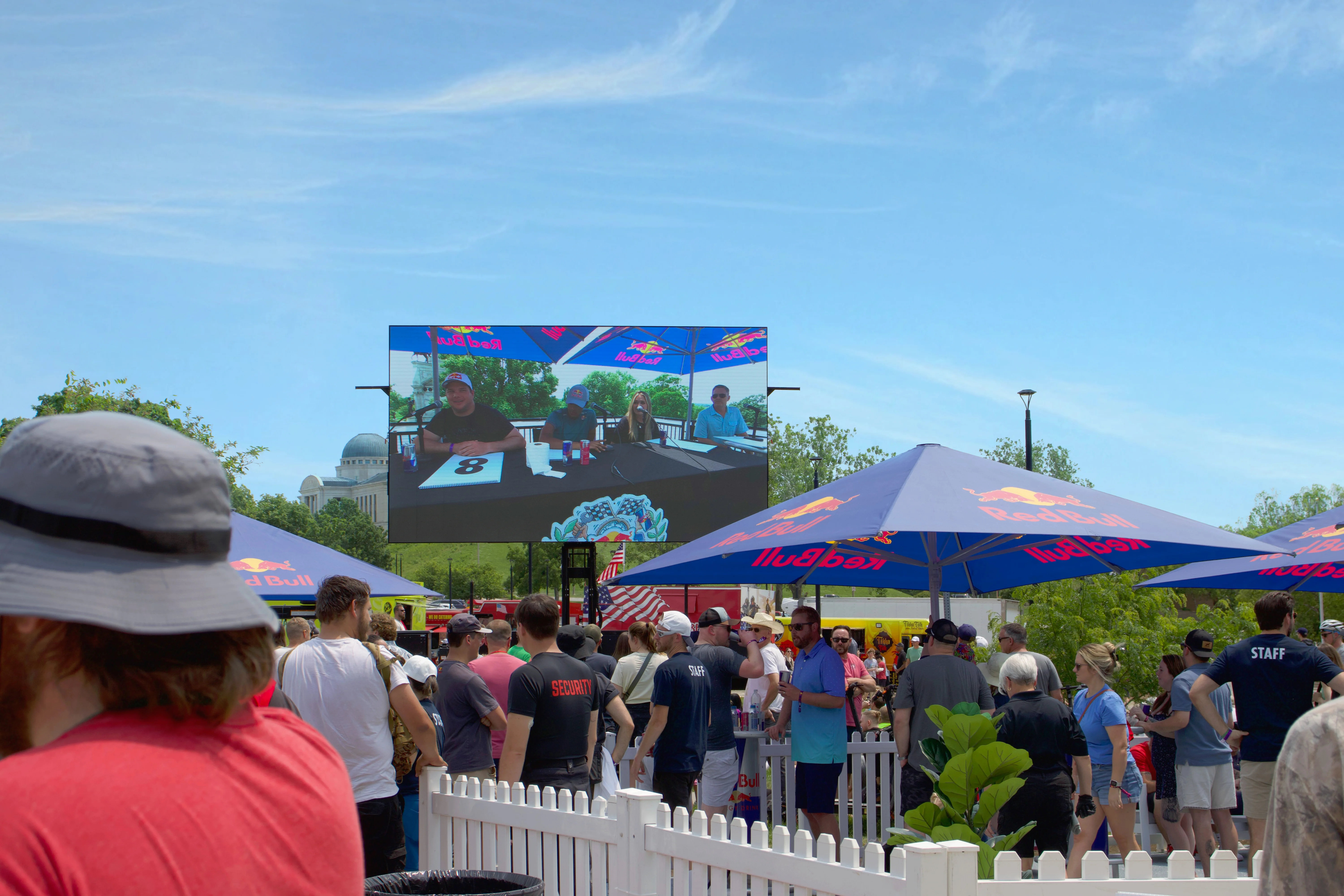 A crowd gathers at the Red Bull Soapbox Race event in Des Moines, watching judges score contestants on a large mobile LED screen under branded umbrellas.