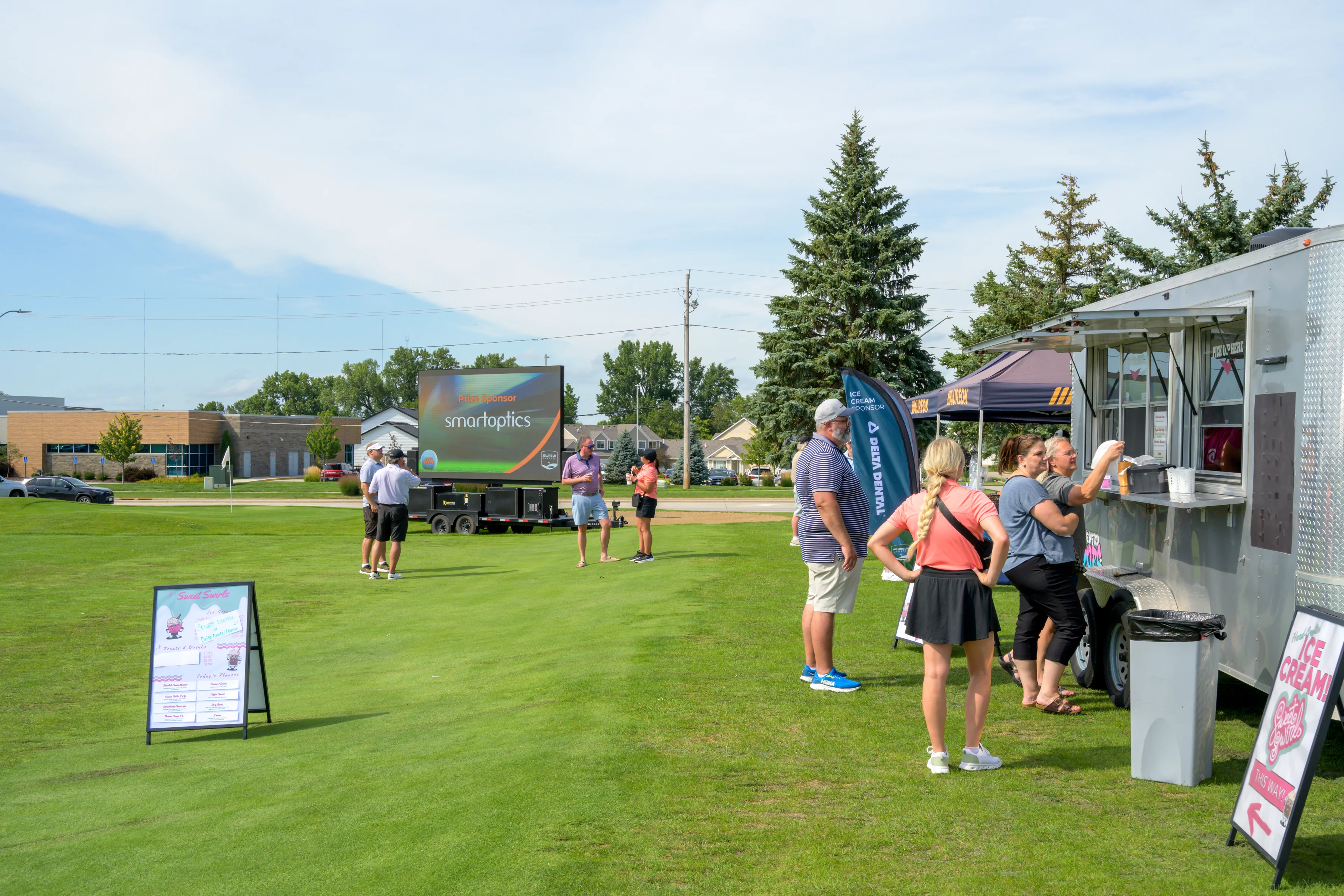 People stand in line at a silver food truck on a golf course, with a large outdoor LED screen nearby showing a sponsor logo.