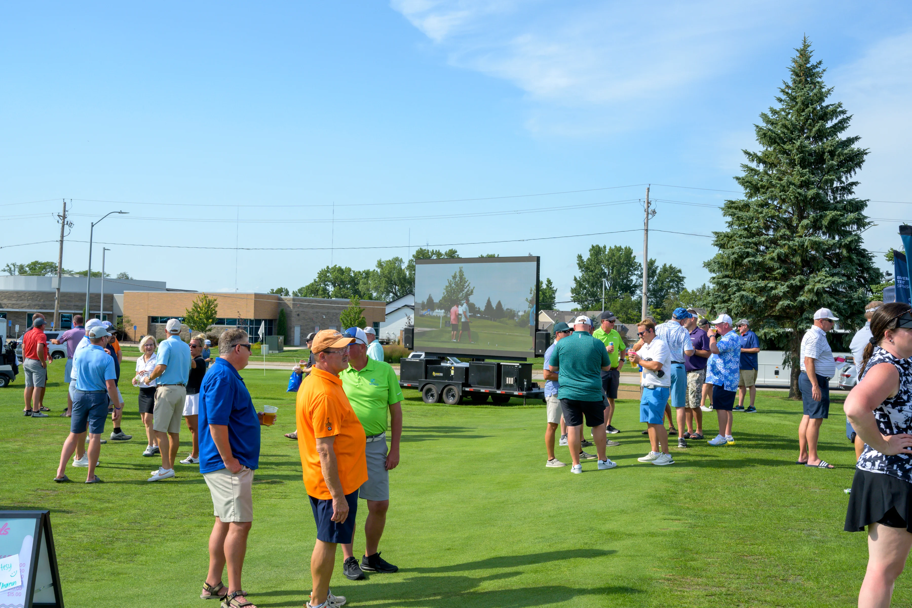 Golfers gather on a green lawn during a sunny day, with a large outdoor LED screen in the background displaying live footage of the event.