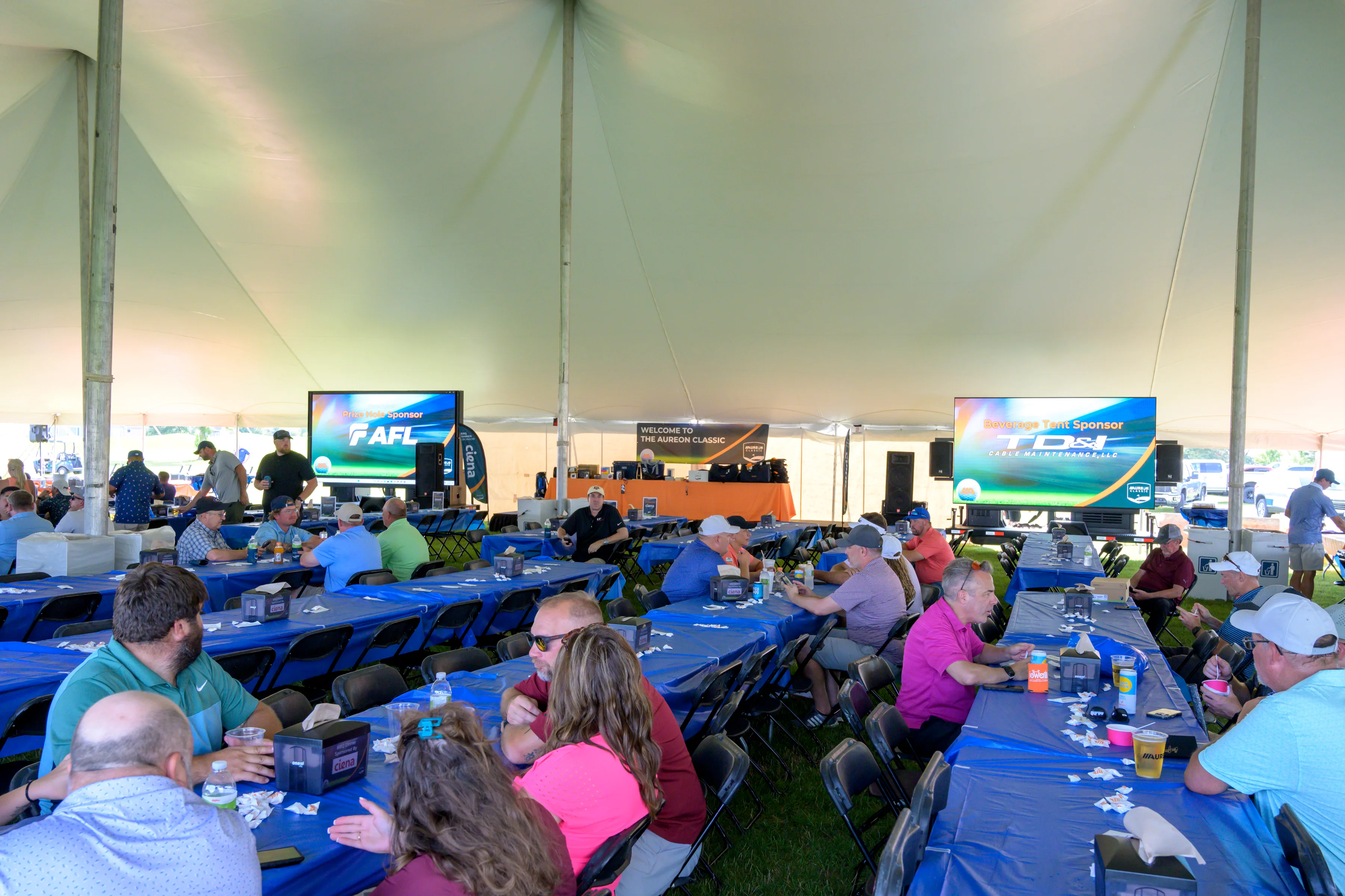 Guests seated under a large white event tent with long tables covered in blue tablecloths, while two indoor LED screens display sponsor messages.