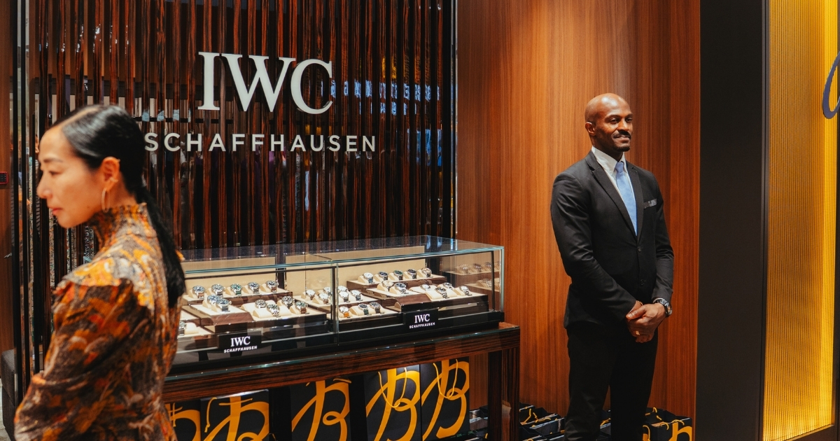Ein Mann in einem schwarzen Anzug steht vor einer IWC Schaffhausen Uhrenvitrine, während eine Frau mit buntem Musterkleid vorbeigeht.