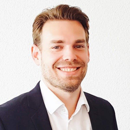 Porträt eines lächelnden Mannes mit braunen Haaren, Anzugjacke und weißem Hemd vor weißer Wand.
