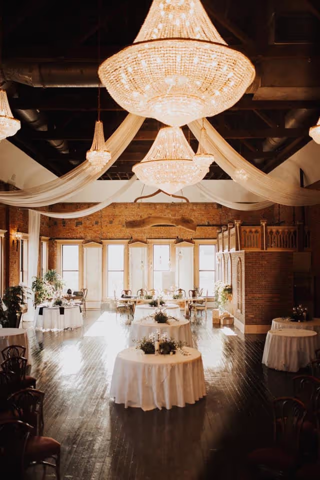The upper room- Elegant event space with exposed brick walls, large chandeliers, white draped fabric across the ceiling, a grand staircase at the center, and round tables covered with white linens, decorated with greenery.