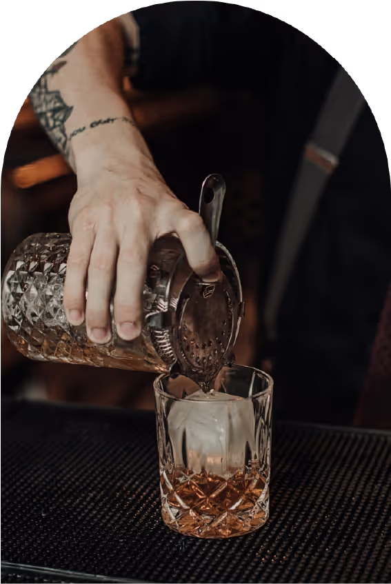 A bartender pours a drink through a strainer into a glass with ice on a bar mat.