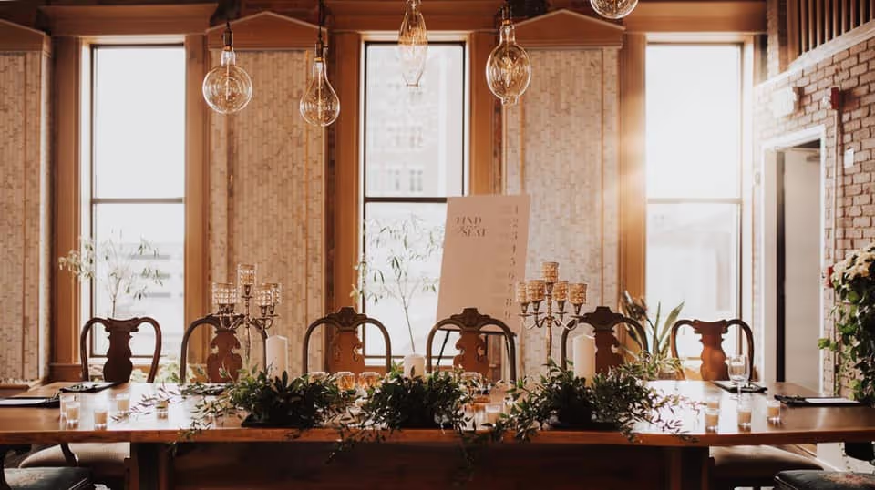 Stylish dining setup with an elegant table, greenery, candles, and hanging lights in a beautifully adorned room with large windows.
