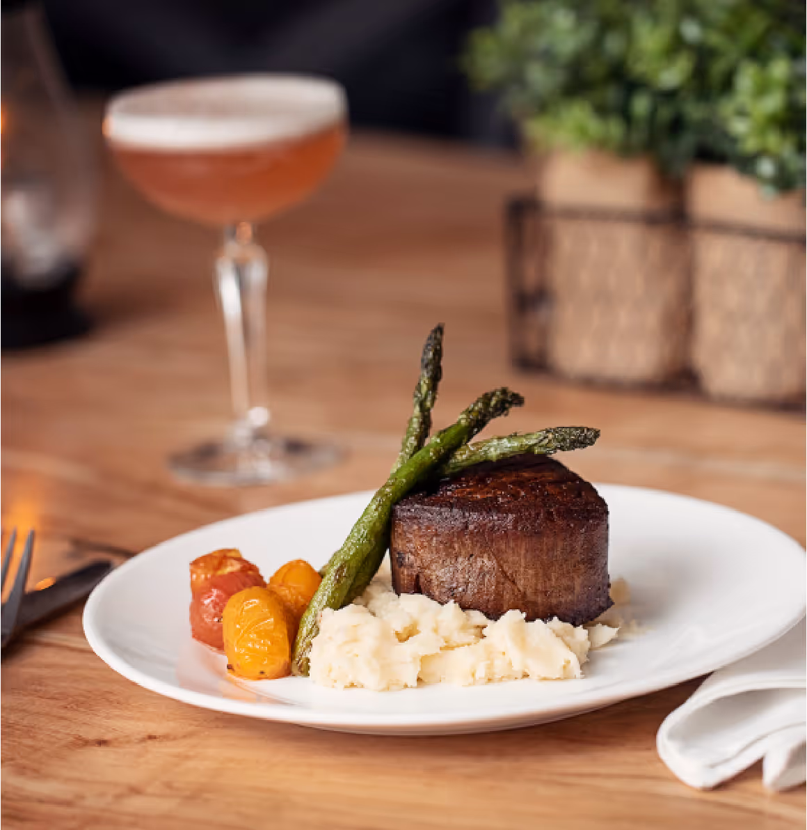 A plated meal with steak, mashed potatoes, asparagus, and tomatoes sits on a wooden table next to a white napkin and a cocktail.