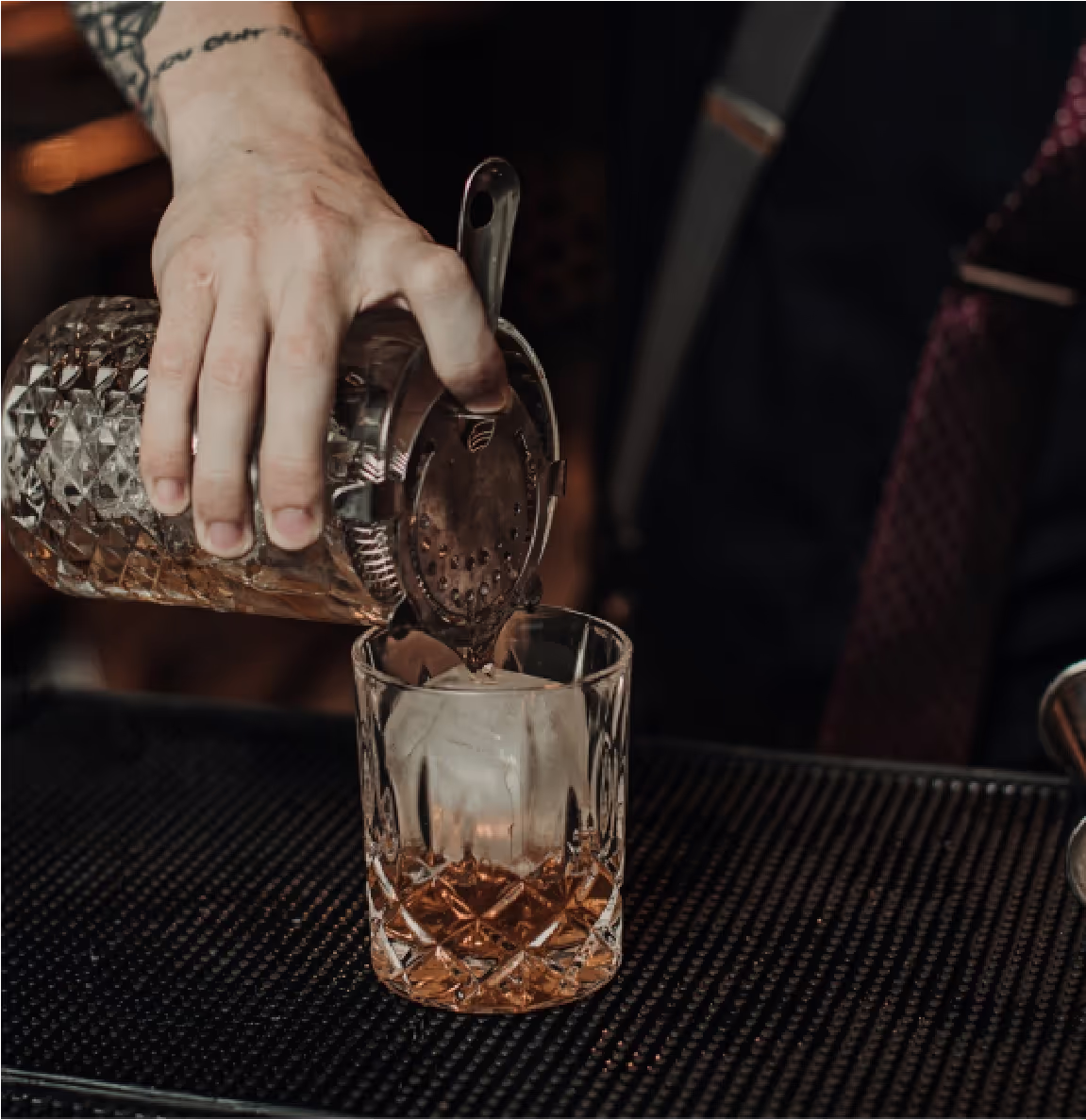 A bartender pours a drink through a strainer into a glass with ice on a bar mat.