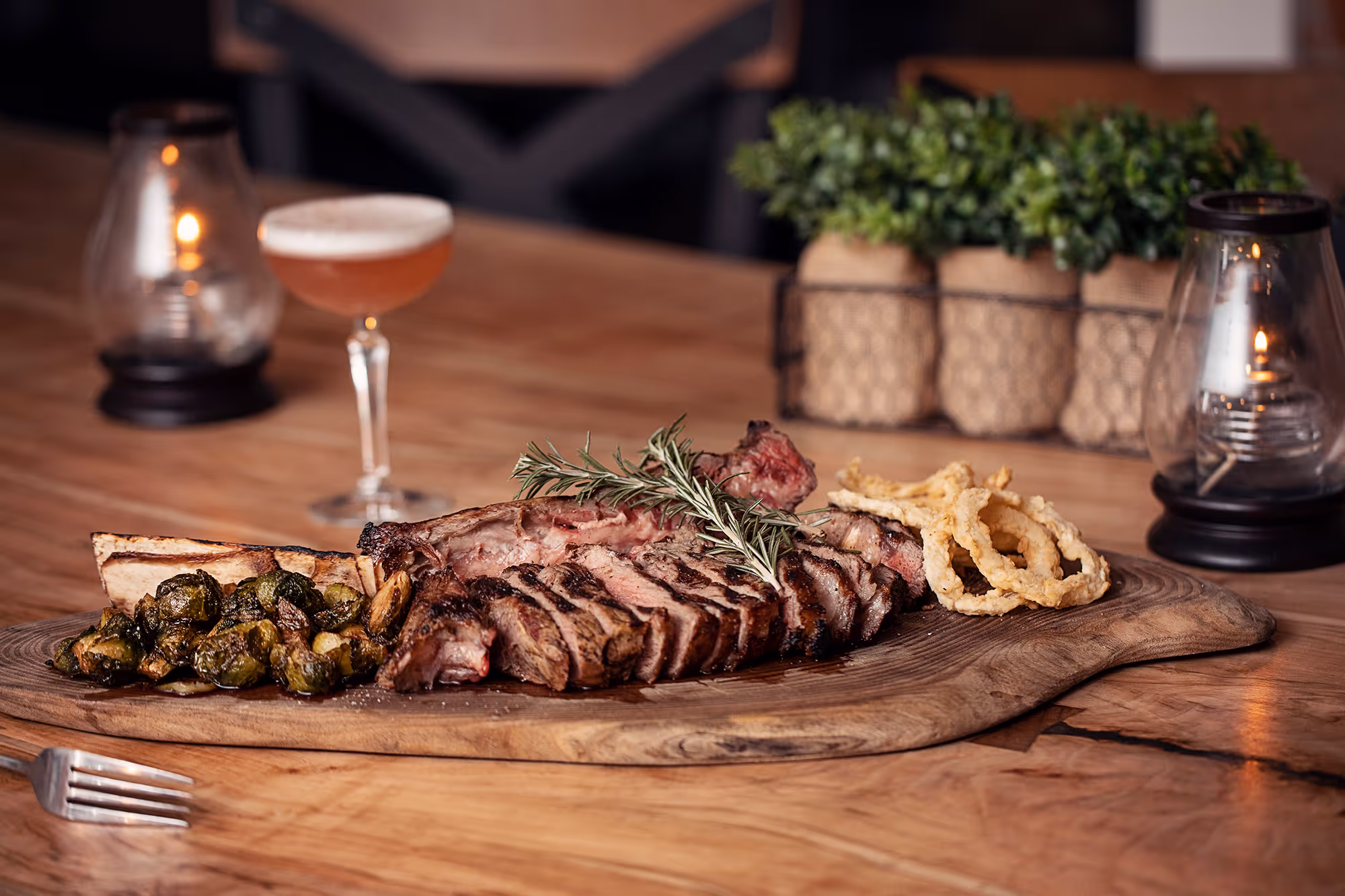 A sliced grilled steak garnished with rosemary, served with roasted Brussels sprouts and a cocktail on a wooden board.