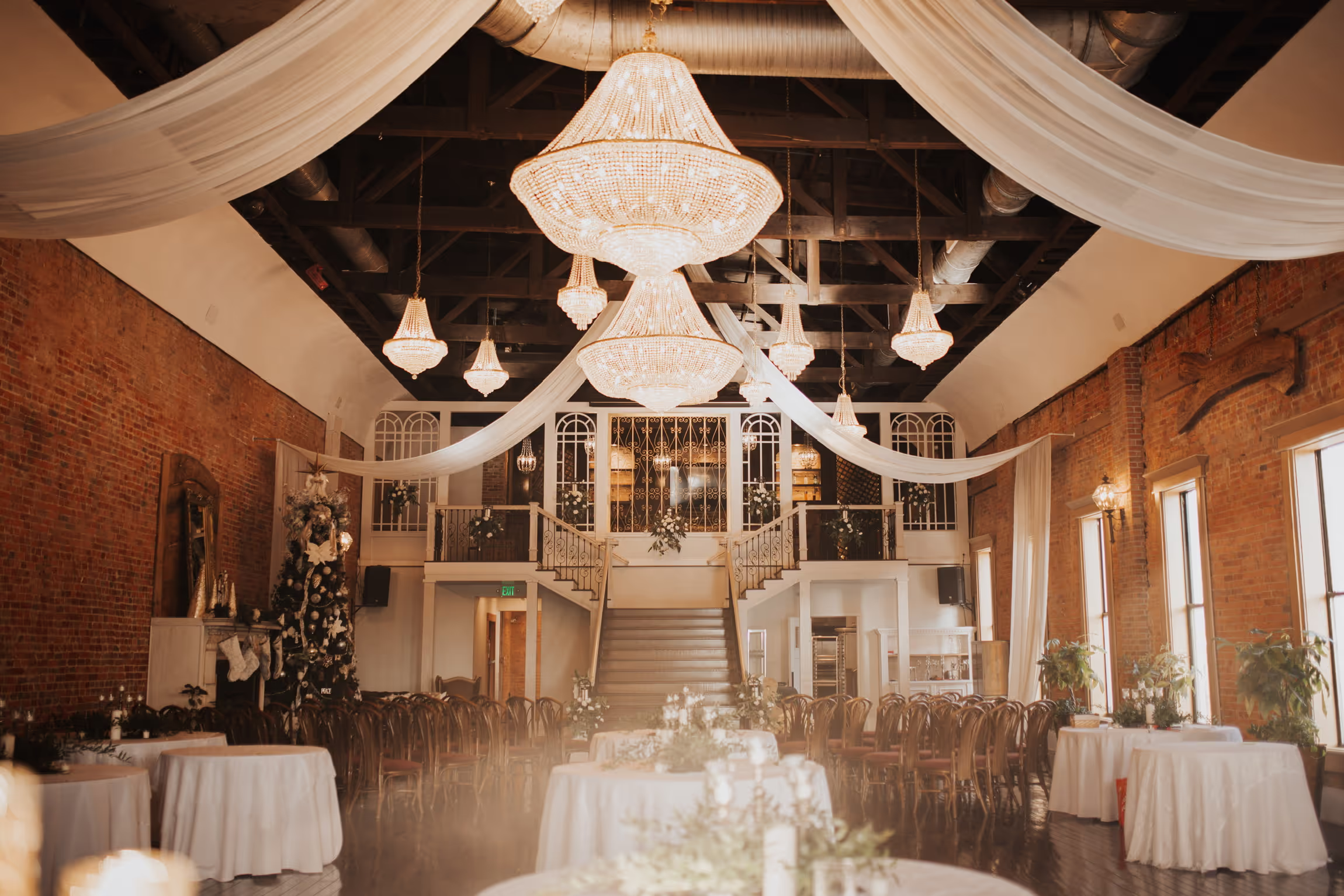 The upper room- Elegant event space with exposed brick walls, large chandeliers, white draped fabric across the ceiling, a grand staircase at the center, and round tables covered with white linens, decorated with greenery.