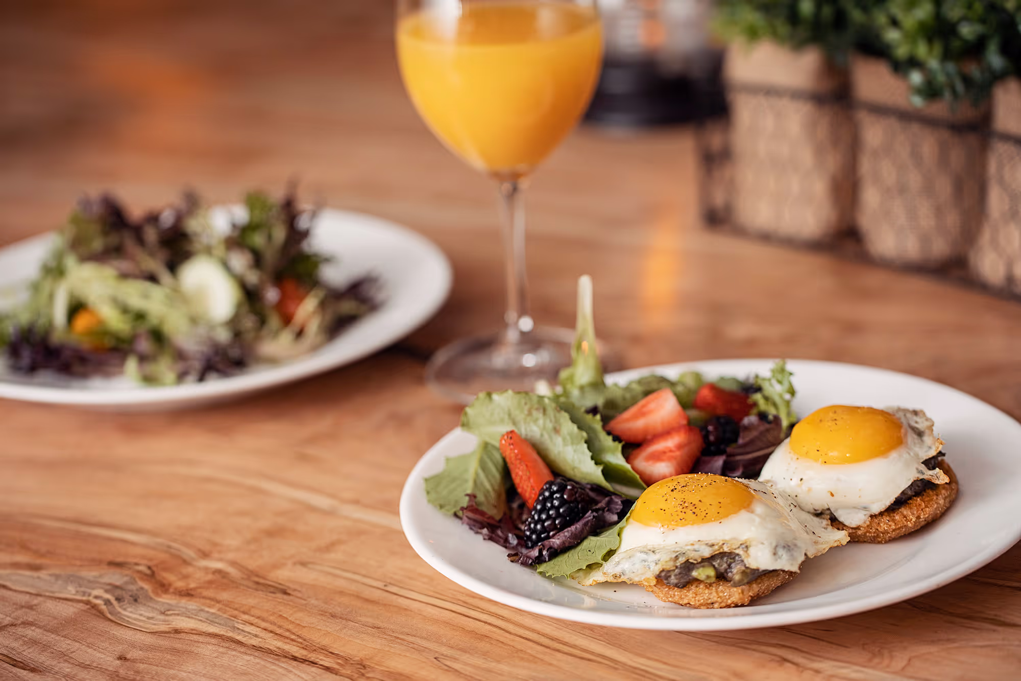 Plate with two fried eggs on toast, surrounded by mixed greens, strawberries, and blackberries, with a glass of orange juice nearby.