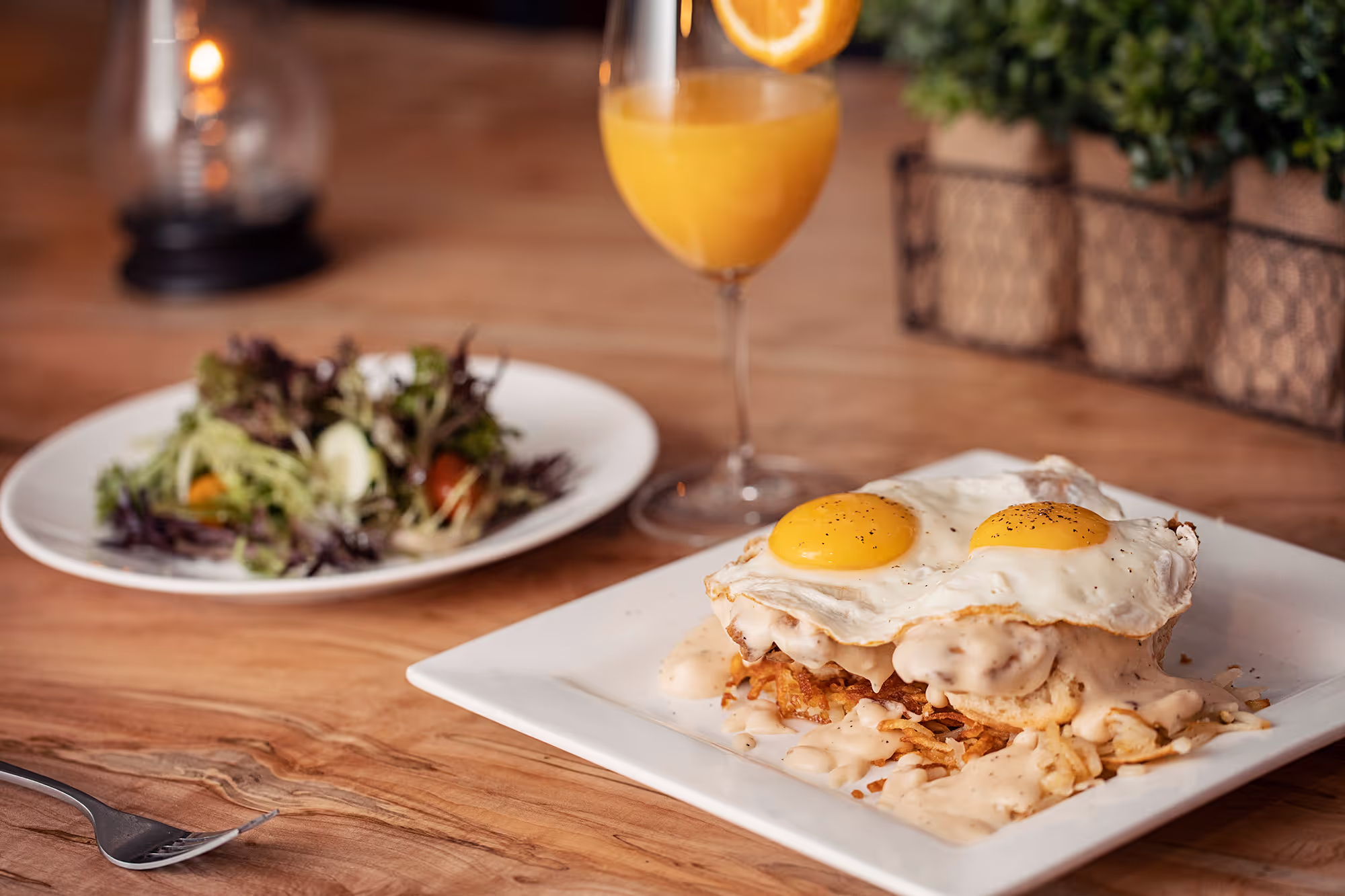 A plate with eggs Benedict, topped with two eggs and creamy sauce, alongside a fresh salad and a mimosa in a cozy setting.