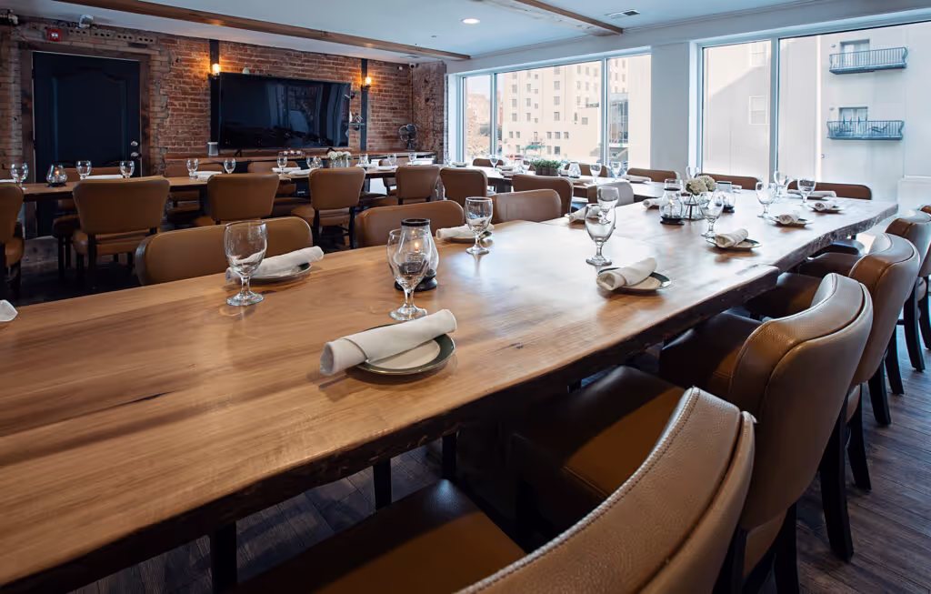 A spacious dining area with a long wooden table set for a meal, surrounded by leather chairs and large windows showcasing the city.