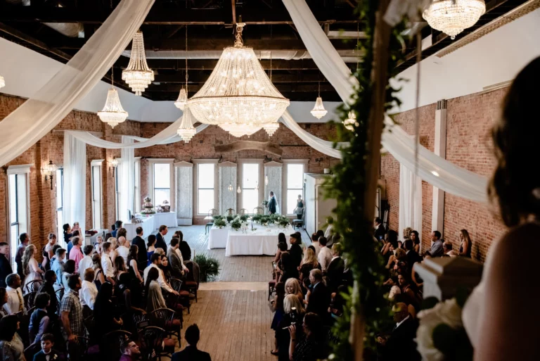 A wedding ceremony in a stylish venue with chandeliers, wooden floors, and guests seated, as the couple stands at the altar.