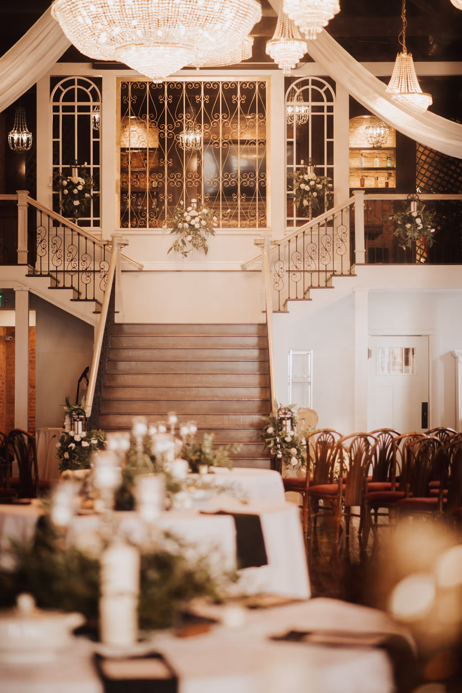 Elegant banquet hall with a grand staircase, adorned with floral arrangements, candles, and chandeliers creating a romantic atmosphere.