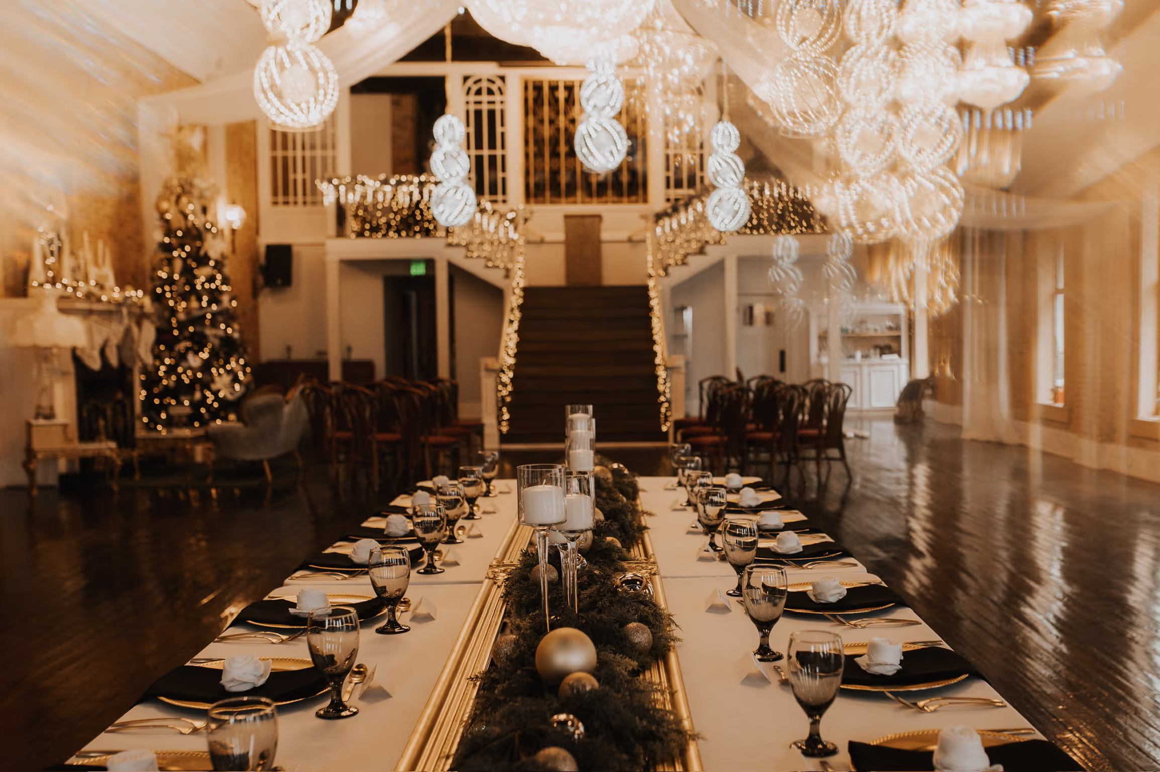 Elegant dining setup with a long table, black and gold decor, candles, and a Christmas tree in a beautifully lit venue.